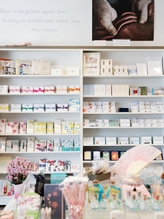 Jenni Bick retail display shelf filled with numerous small gift boxes, cards, and packaged items, with a decorative wall quote above and a fan in the foreground.